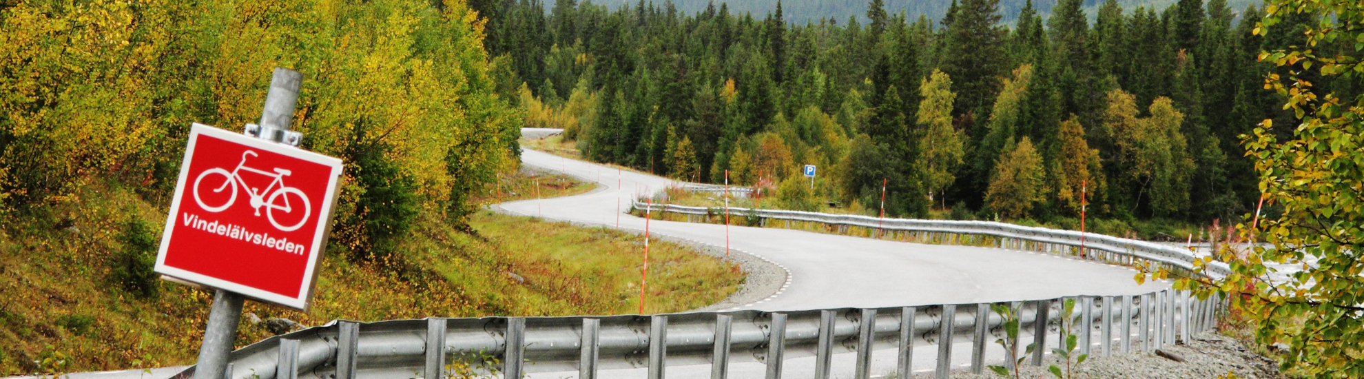 En slingrig väg omgiven av skog och en skylt i förgrunden som det står Vindelälvsleden på.